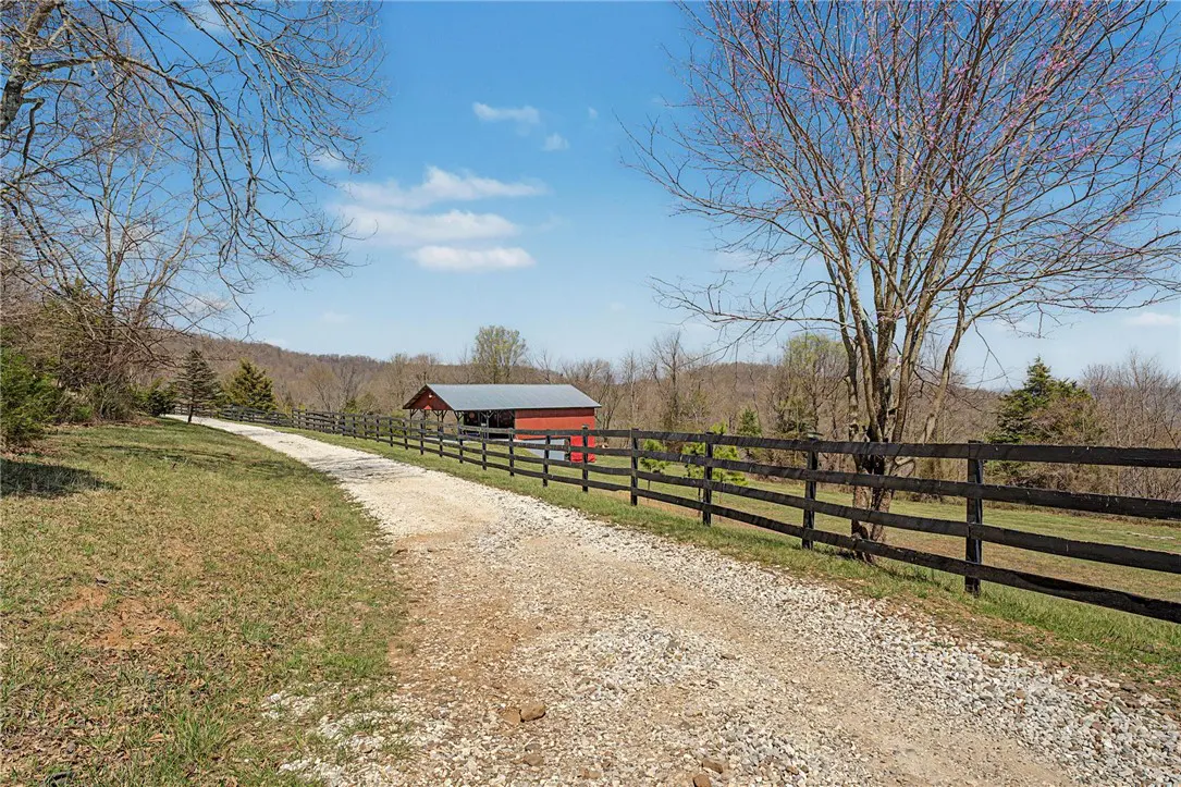 Gravel driveway leading to a red barn with a black fence on a sunny day.