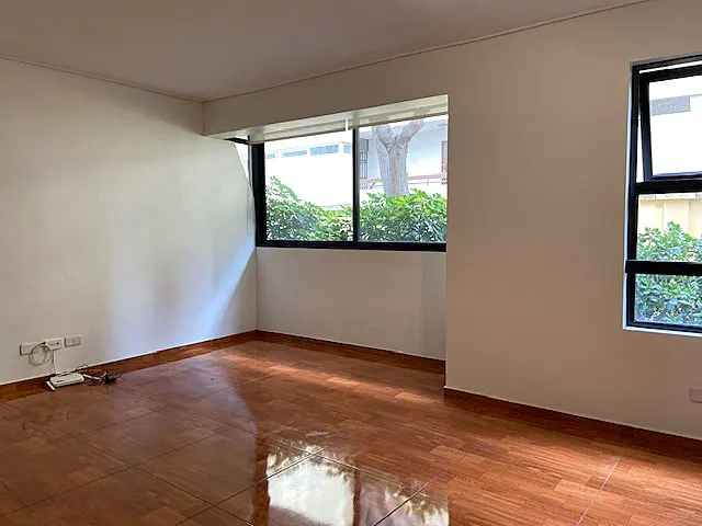 Empty room with light walls and shiny wood floors. Two windows with black frames look out onto greenery.