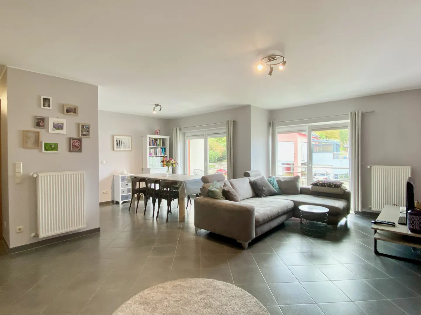 Open-concept living and dining area with gray walls, tile floors, and natural light. A gray sectional sofa faces a TV stand. A dining table with chairs sits near a bookshelf.