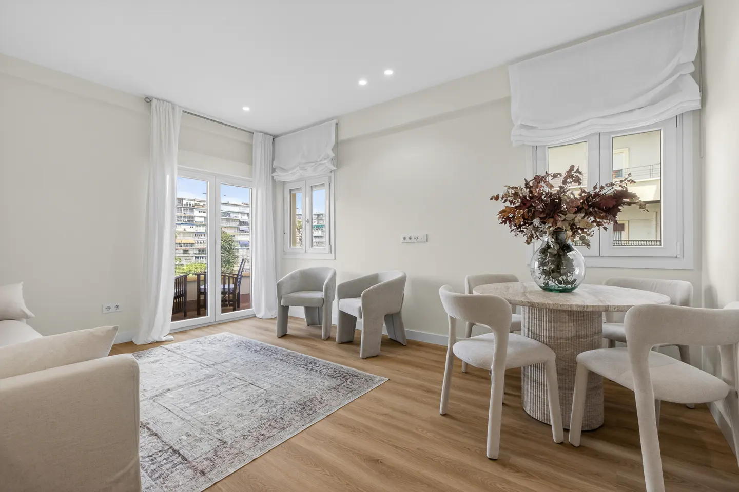 Bright living room with wood floors, white walls, and neutral furniture. A round table with a floral arrangement sits near a window.