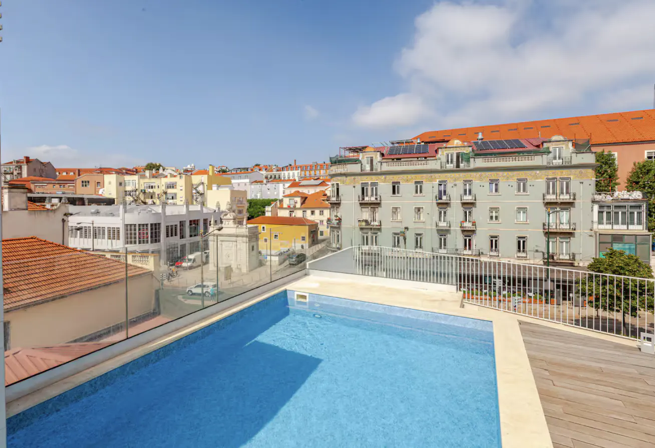 Rooftop pool with blue tiles and wooden deck overlooking a European city with orange rooftops under a blue sky.