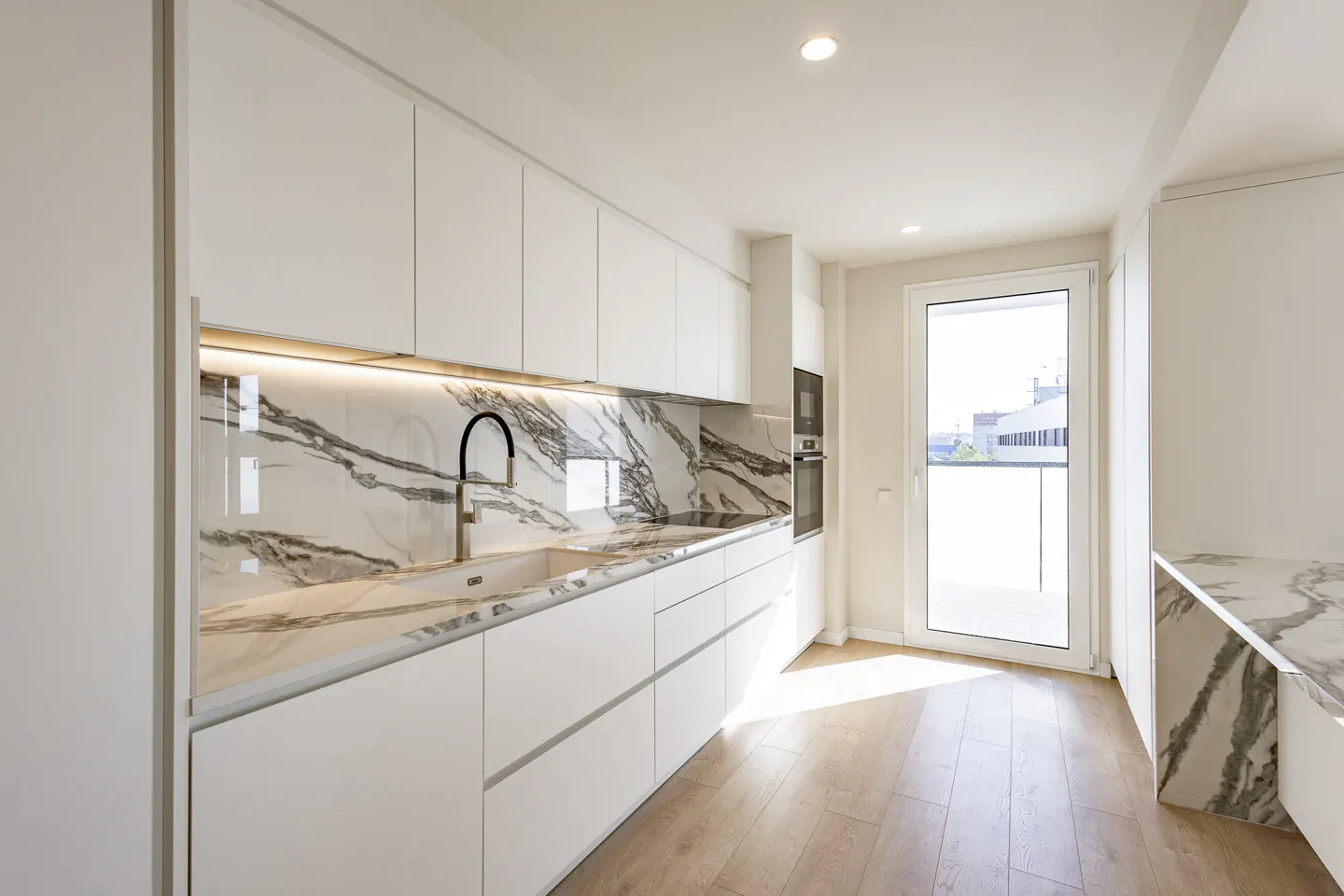 Bright, modern kitchen with white cabinets, marble backsplash and countertops, and wood floors. A door leads to a balcony.
