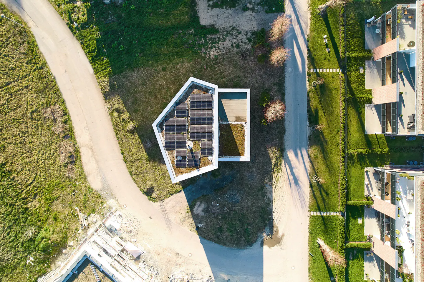 Aerial view of a modern white house with solar panels and a green roof, surrounded by green grass and a road.