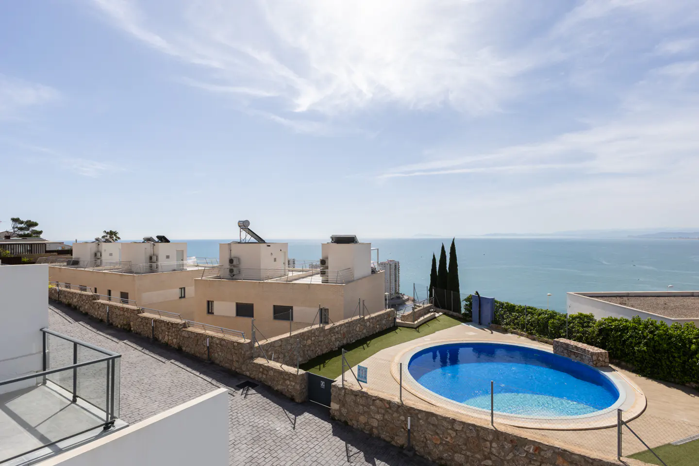 View of modern beige buildings with a blue pool, stone walls, and ocean backdrop under a bright sky.