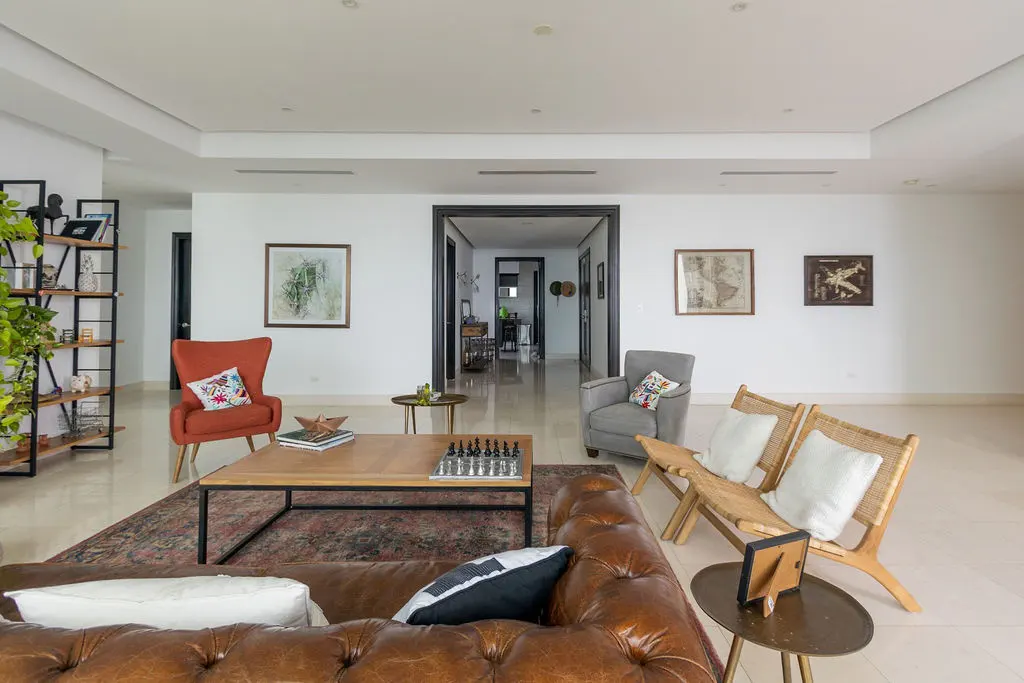 Bright living room with a brown leather couch, chess set on a wood coffee table, and two chairs. Artwork hangs on the white walls.