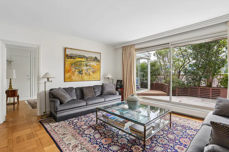 Living room with gray sofa, glass coffee table, and a large painting. Sliding glass doors open to a patio with greenery.