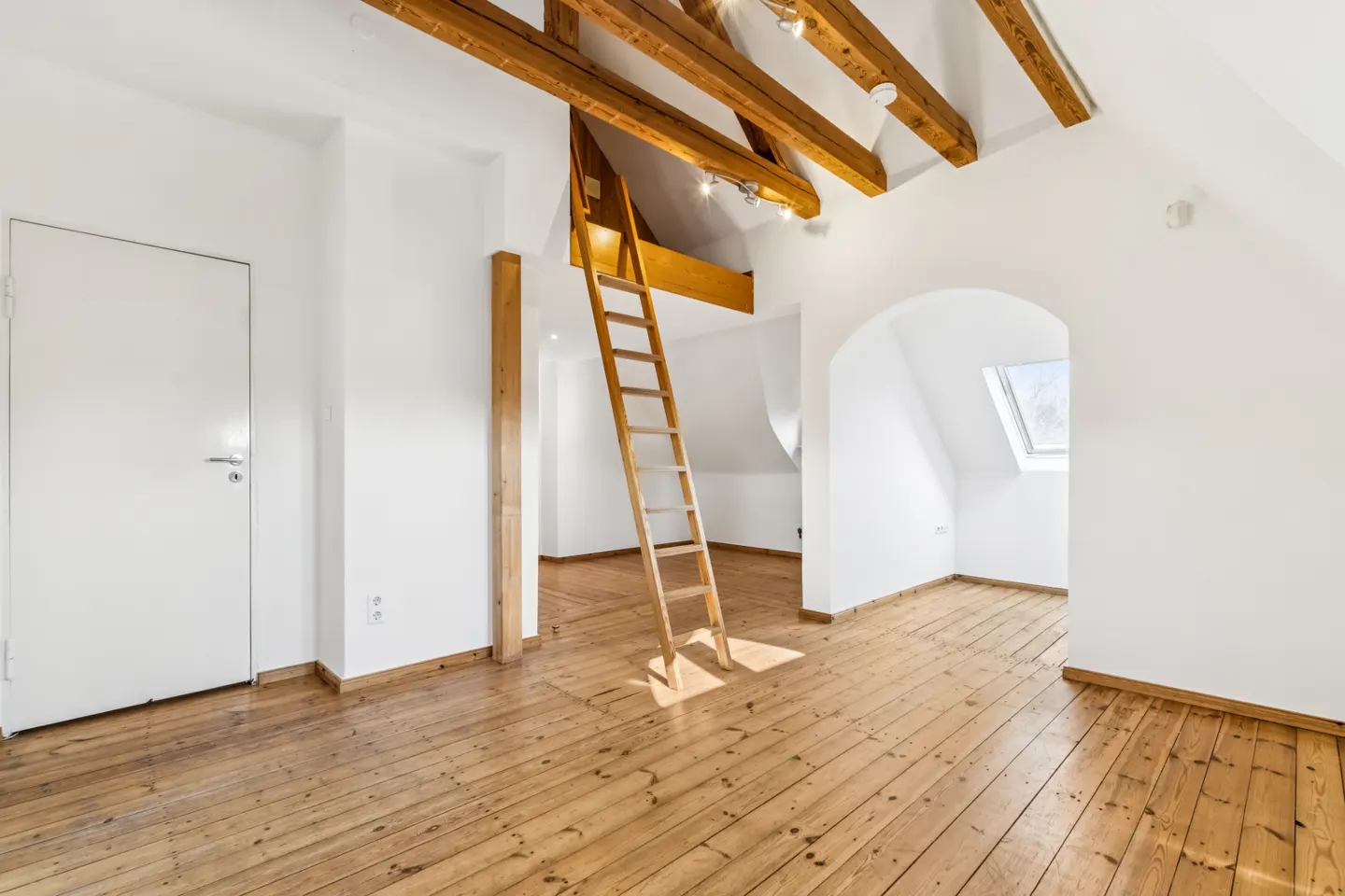 Attic room with wooden floors, white walls, and exposed beams. A ladder leads to an upper level. Arched doorway and skylight visible.