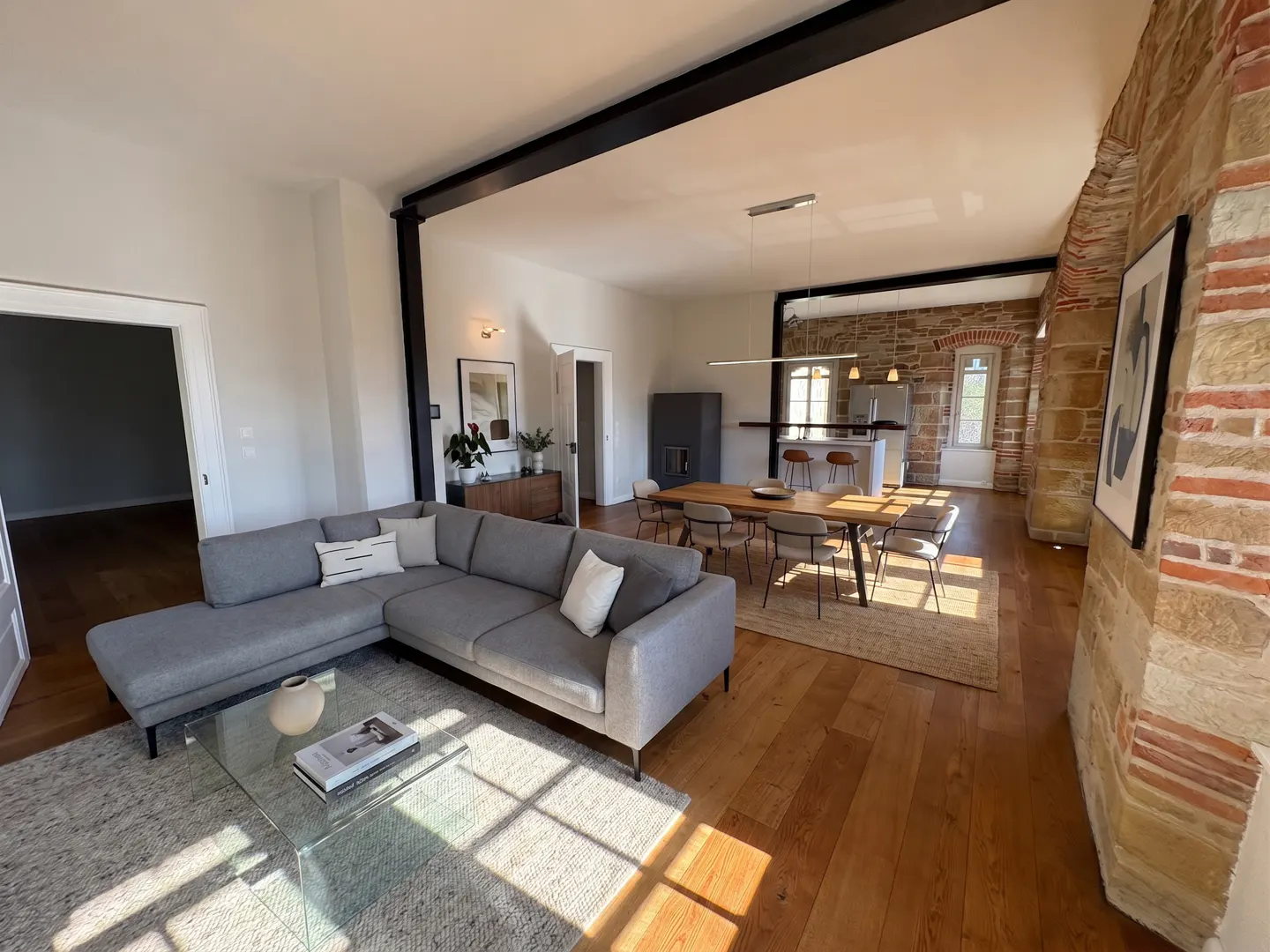 Open-concept living space with a gray sectional sofa, glass coffee table, and wood floors. Dining table and kitchen in the background.