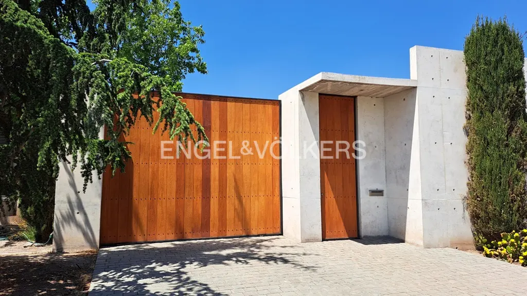 Modern concrete wall with a large wooden gate and matching door, set against a clear blue sky and lush green trees.