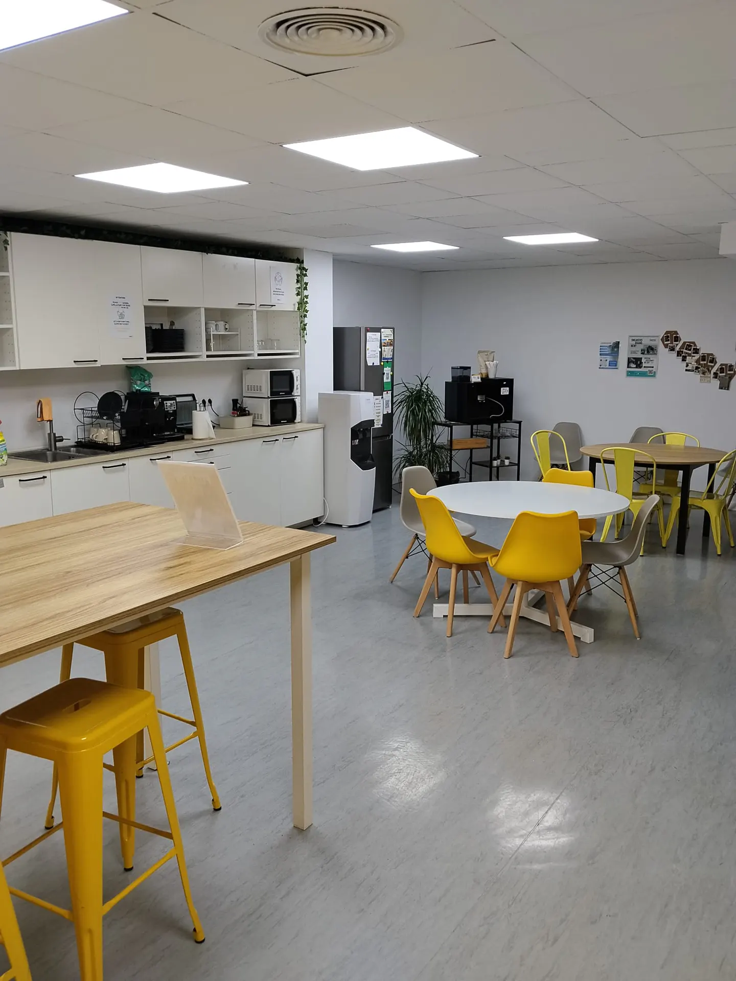 Bright office breakroom with white cabinets, a wooden table with yellow stools, and round tables with yellow chairs.