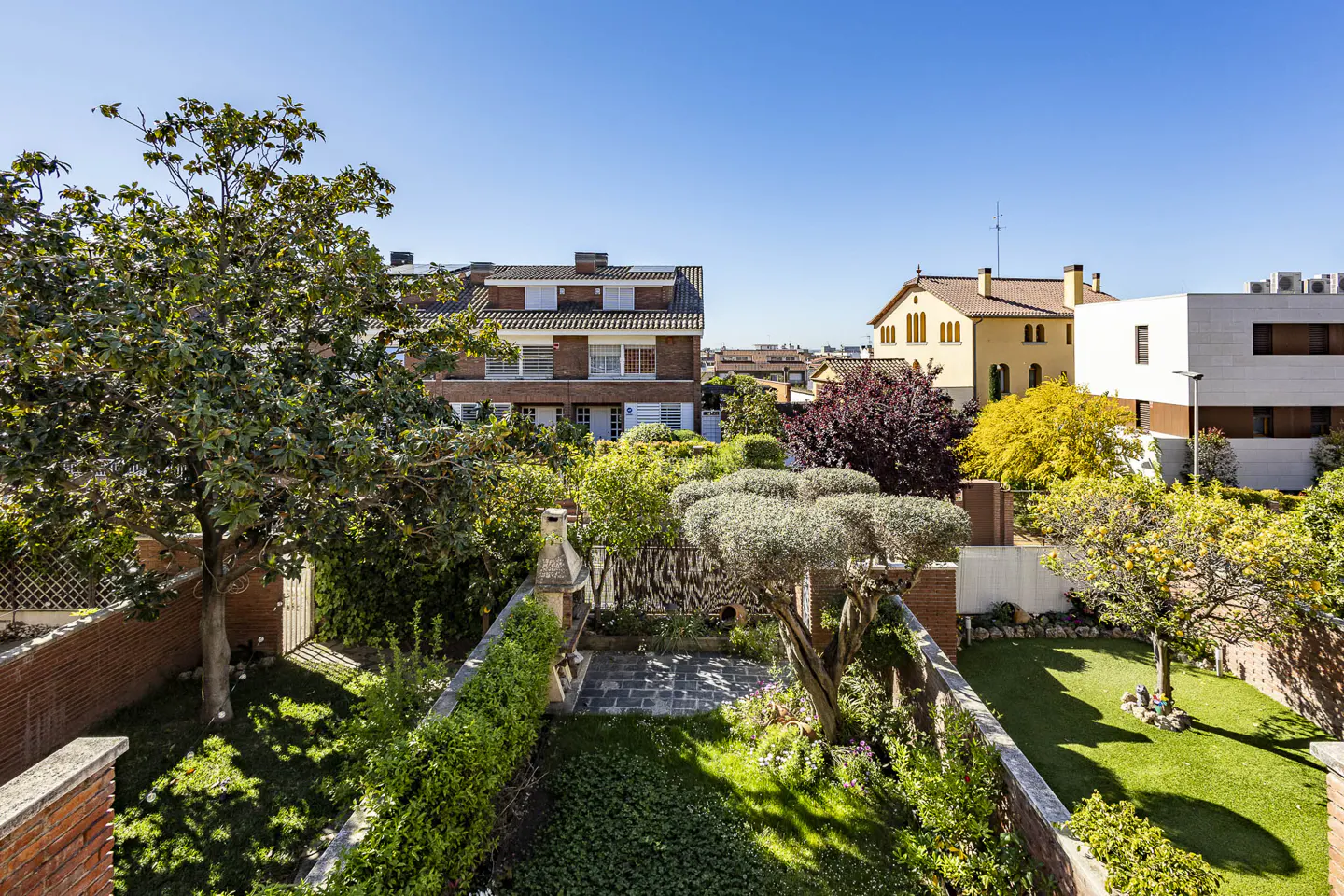 A high angle view of a backyard with green grass, trees, and a brick wall, with houses in the background under a blue sky.