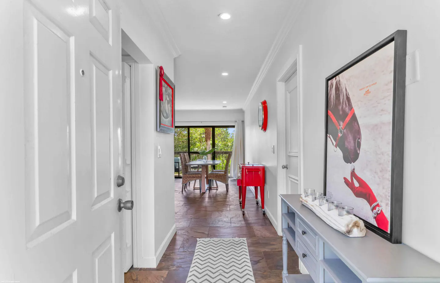 A bright hallway with a white door, gray walls, and a brown tile floor. A gray console table with a horse art piece is on the right. A dining area is visible at the end.