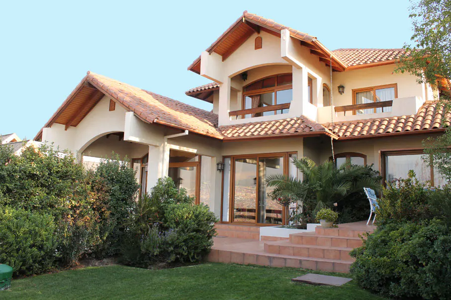 Two-story house with a red tile roof, beige stucco walls, and a green lawn. Bushes and trees surround the house.