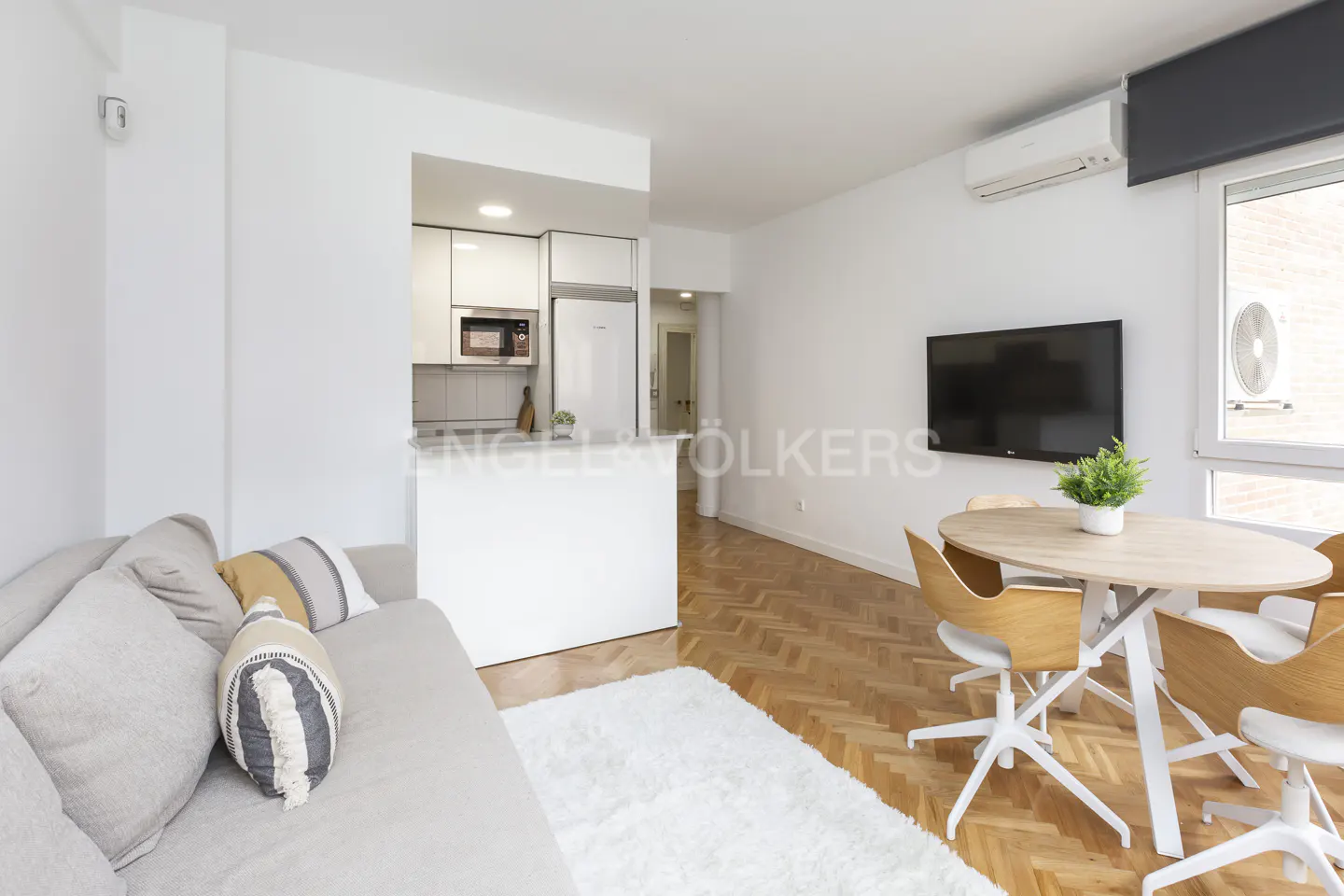 Bright, modern apartment interior with a beige sofa, white rug, and a round table with four chairs. A small kitchen is visible in the background.