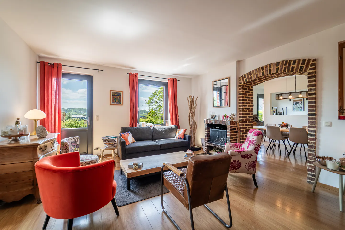Bright living room with hardwood floors, gray sofa, orange and floral armchairs, and a brick archway leading to the dining area.