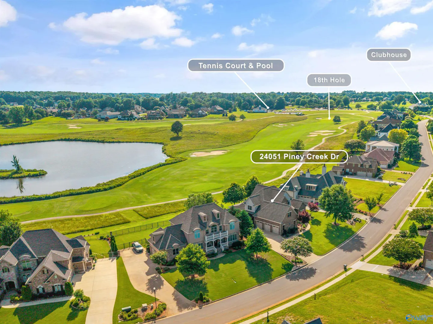 Aerial view of a residential street overlooking a golf course, pond, tennis court, pool, and clubhouse on a sunny day.