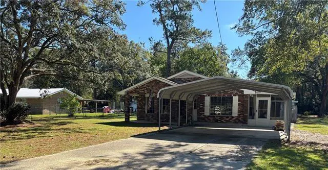 A single-story brick house with a metal carport and a concrete driveway on a sunny day. Trees surround the house.