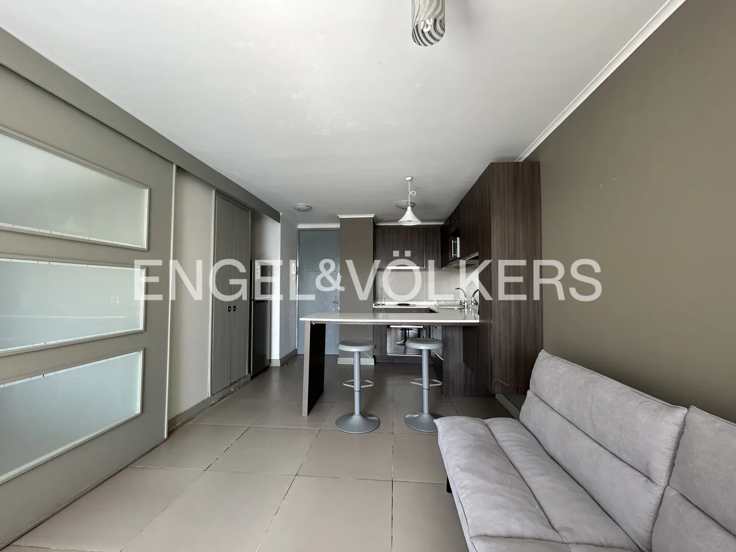 A modern apartment interior with a gray sofa, kitchen island with stools, and dark wood cabinets.