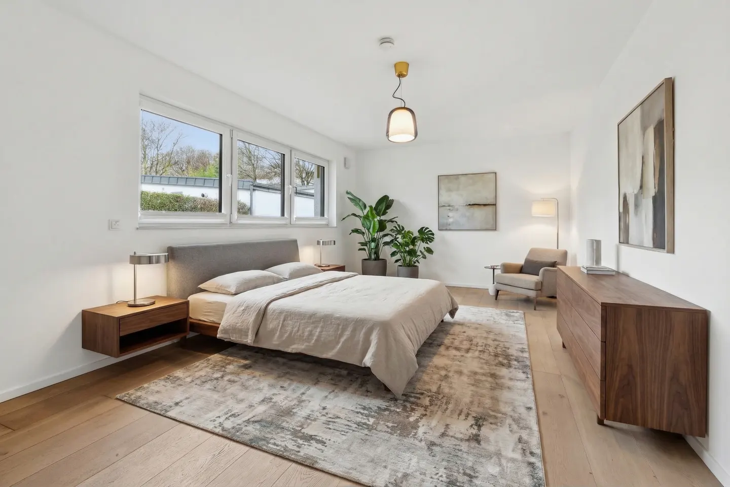 Bright bedroom with a gray bed, wood floors, and white walls. A large window lets in natural light. Artwork and plants add to the decor.