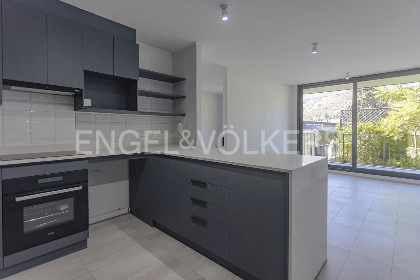 A modern kitchen with gray cabinets, white countertops, and stainless steel appliances. A large window overlooks a garden.