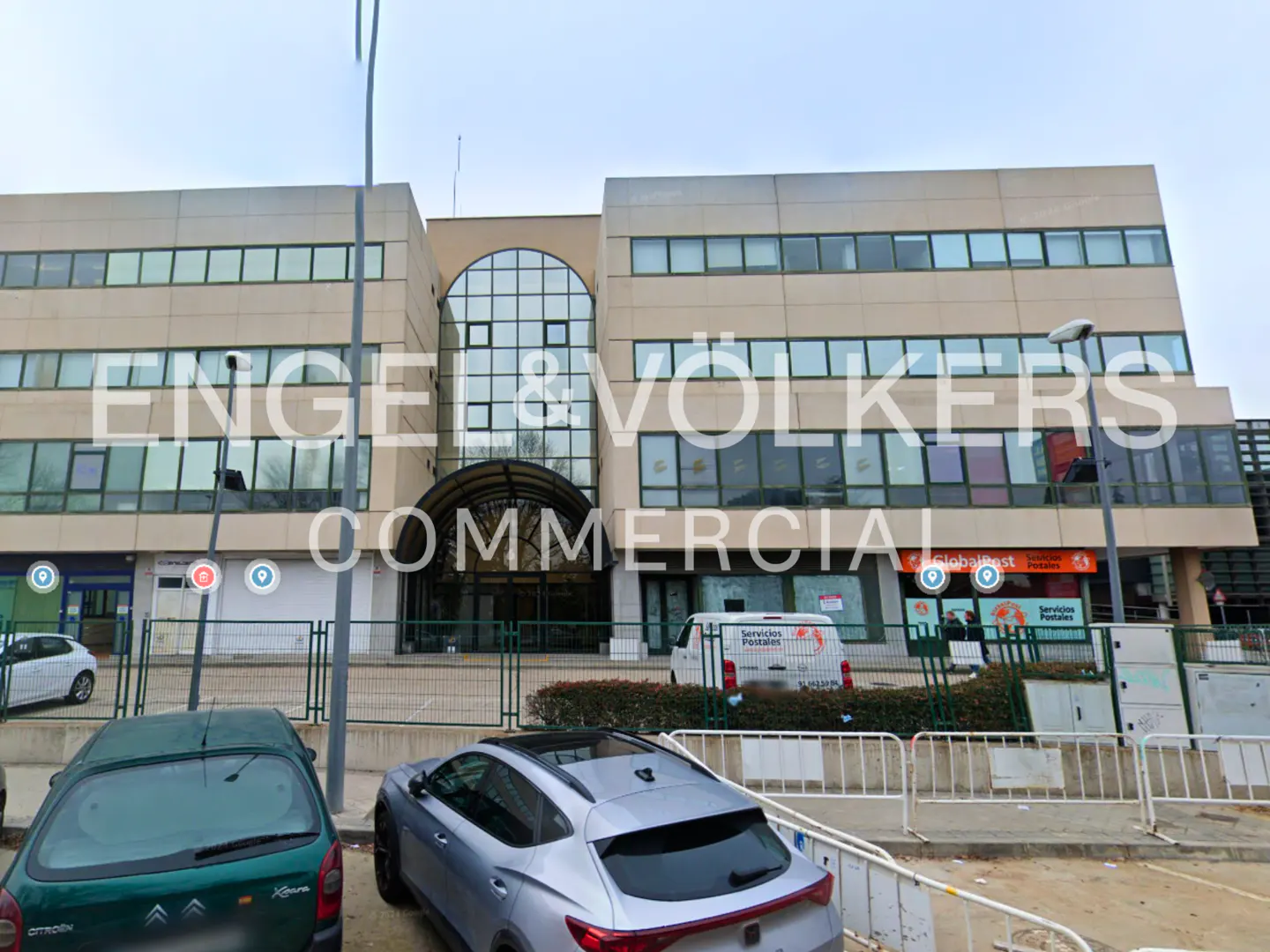 Exterior of a beige, multi-story Engel & Völkers Commercial building with a glass arched entrance and parked cars in front.