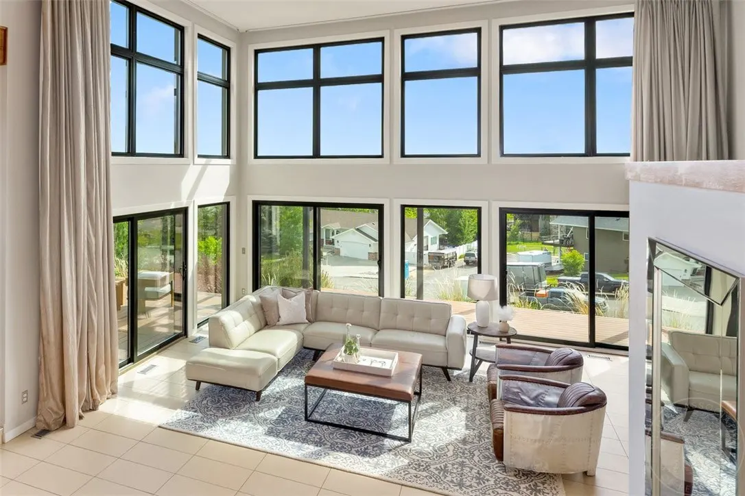 Modern living room with high ceilings, floor-to-ceiling windows, a white sectional sofa, and two leather armchairs.