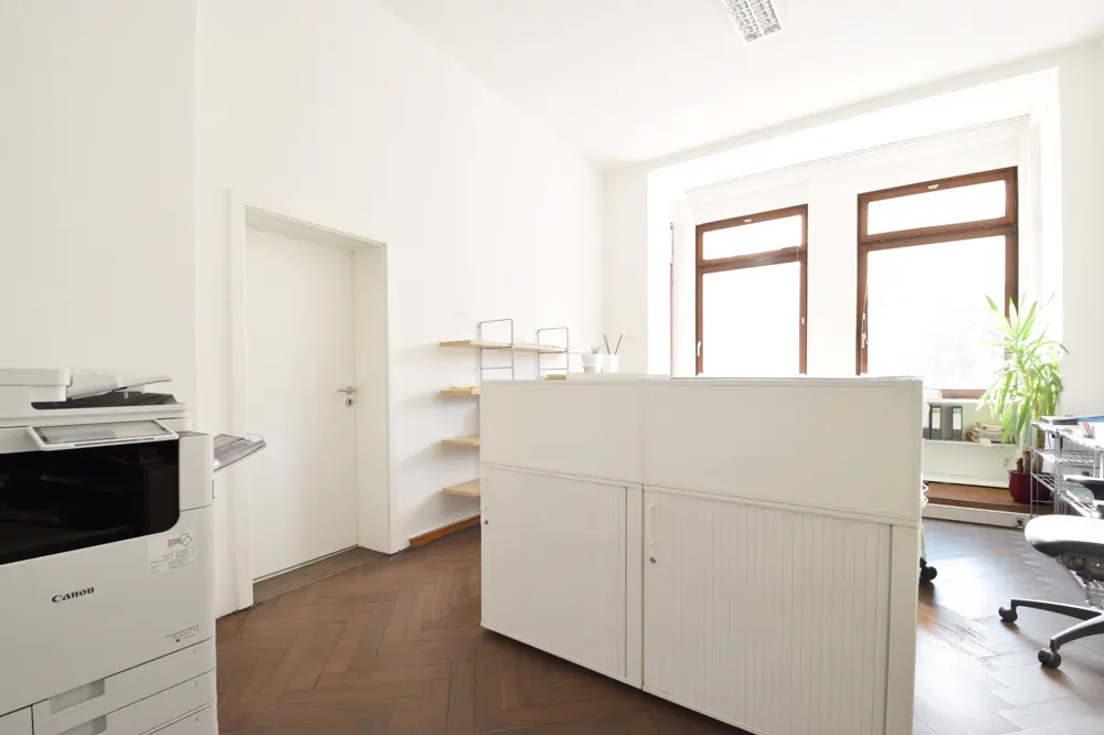 Bright office space with white walls, wood floors, and large windows. A Canon printer sits near a closed white door. A white reception desk is in the center.