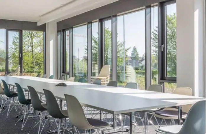 Bright conference room with a long white table and colorful chairs. Large windows overlook green trees.