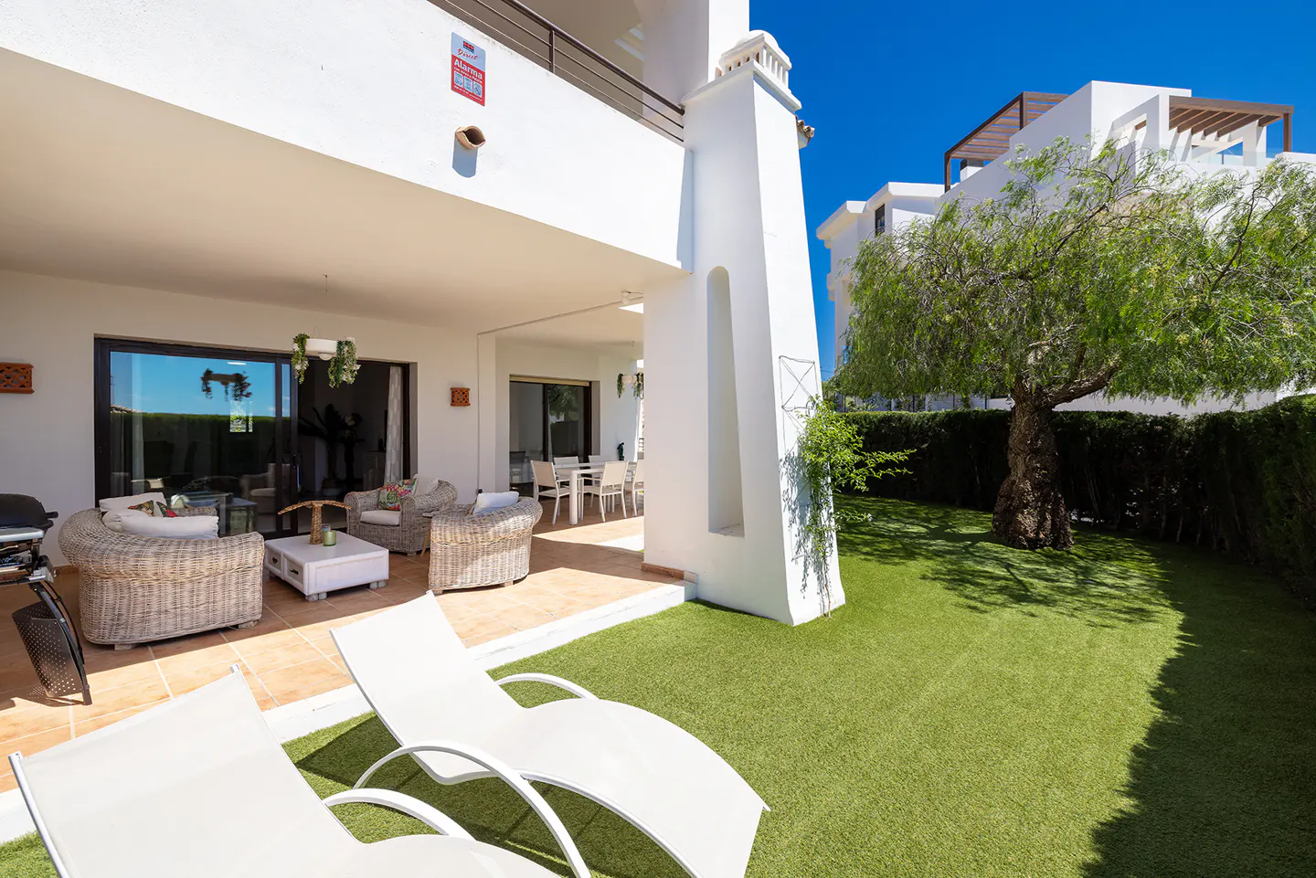 Outdoor patio with white lounge chairs, wicker furniture, and green lawn. Sunny day, blue sky.