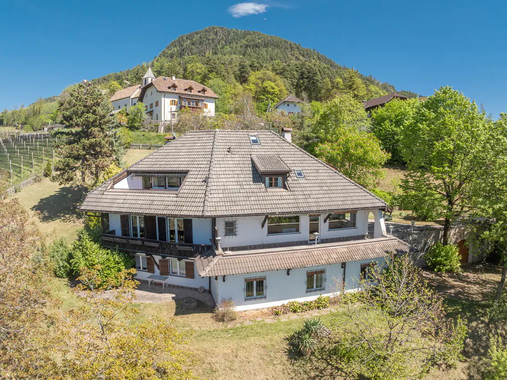 Two-story white house with a gray tiled roof, surrounded by green trees and a mountain in the background on a sunny day.