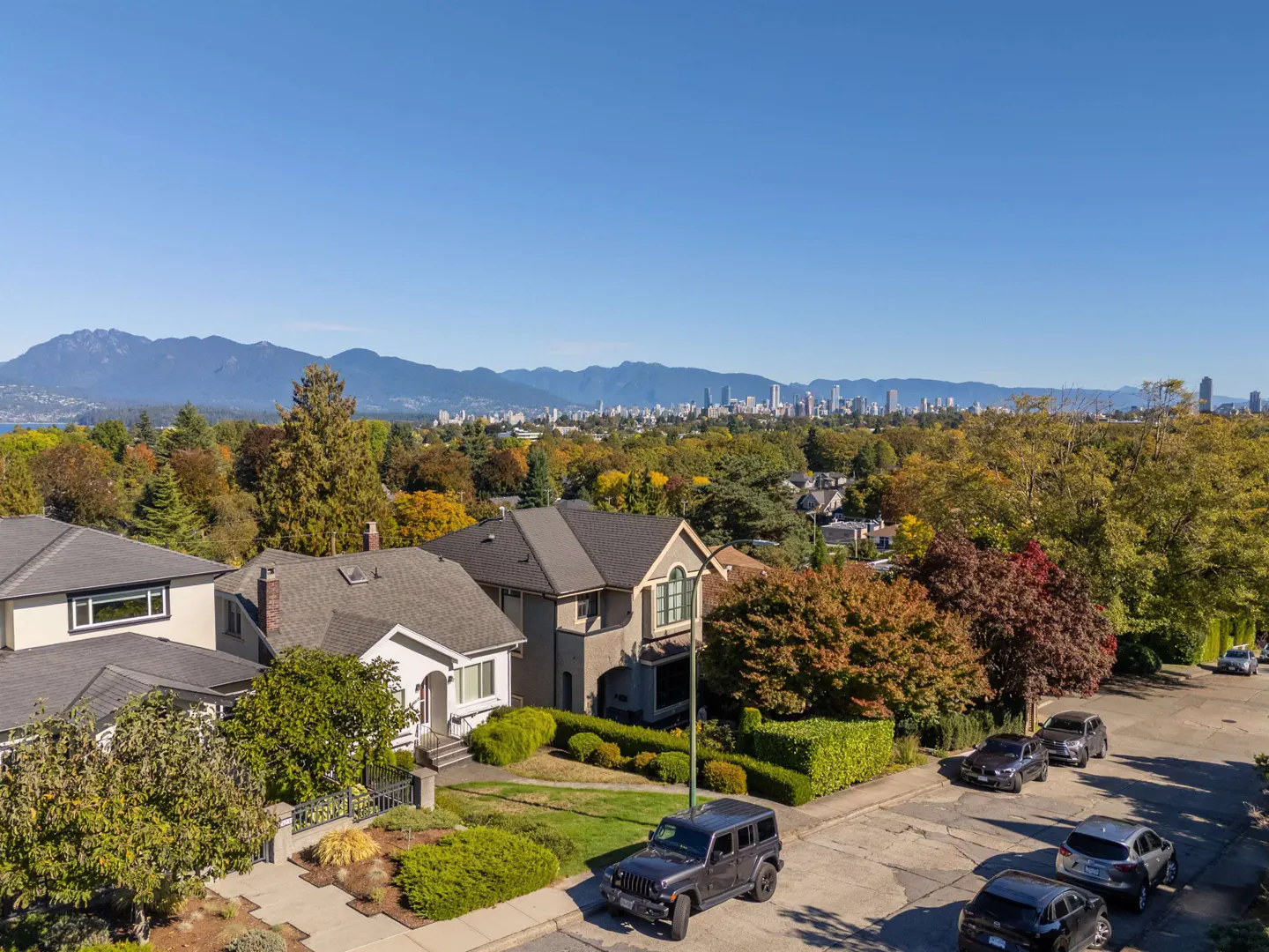 Residential street view with houses, cars, and trees in autumn colors. Distant mountains and city skyline under a clear blue sky.