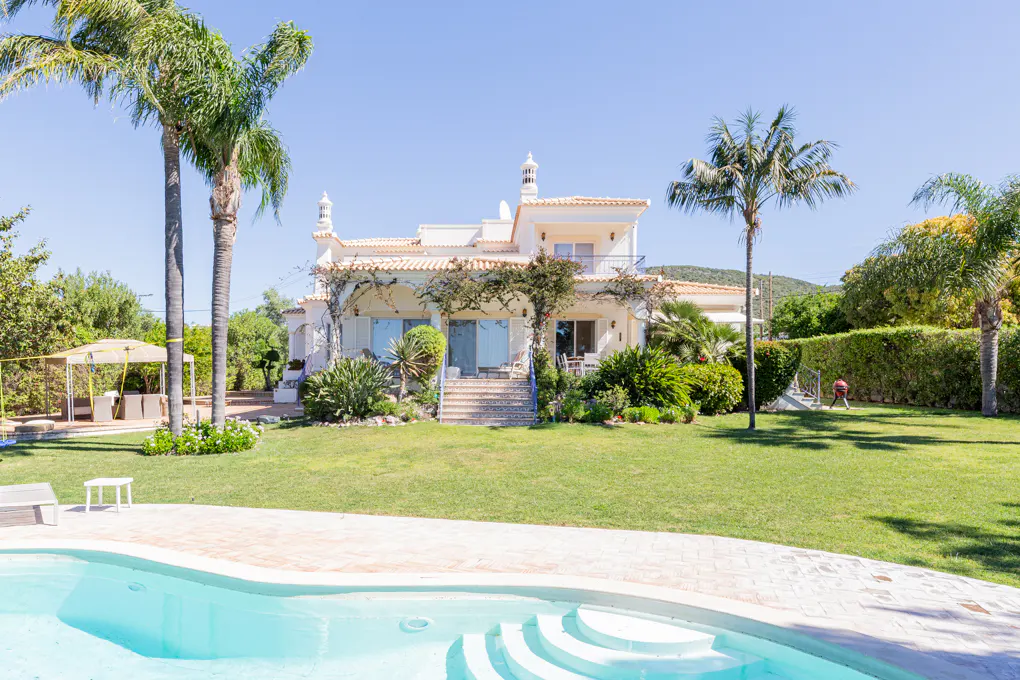Two-story white villa with a pool, lawn, and palm trees under a clear blue sky.