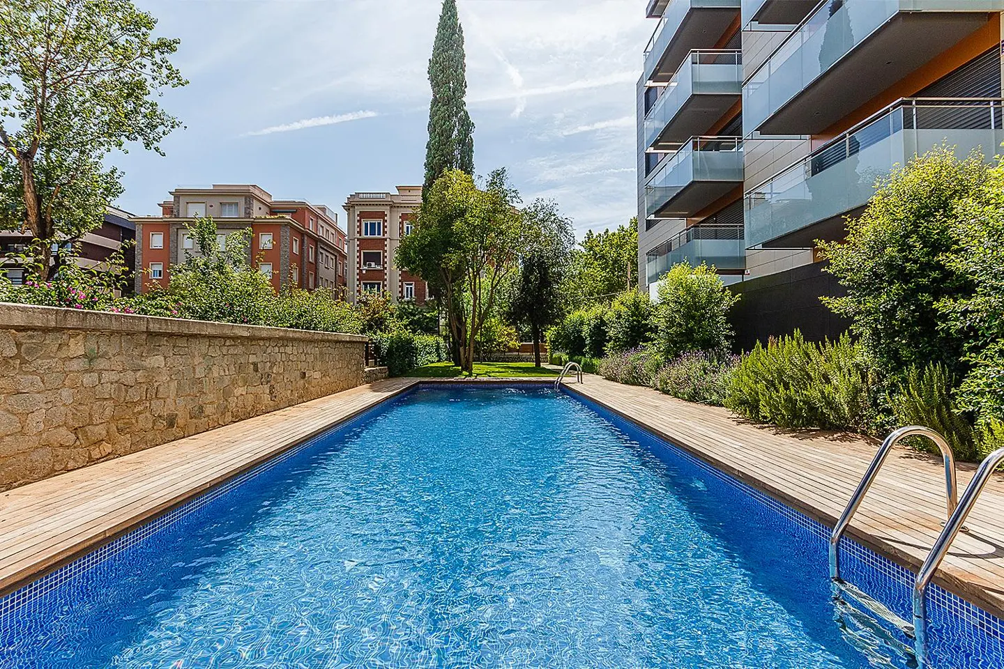 Outdoor pool with blue tile, wooden deck, and metal ladder. Buildings and greenery in the background under a blue sky.