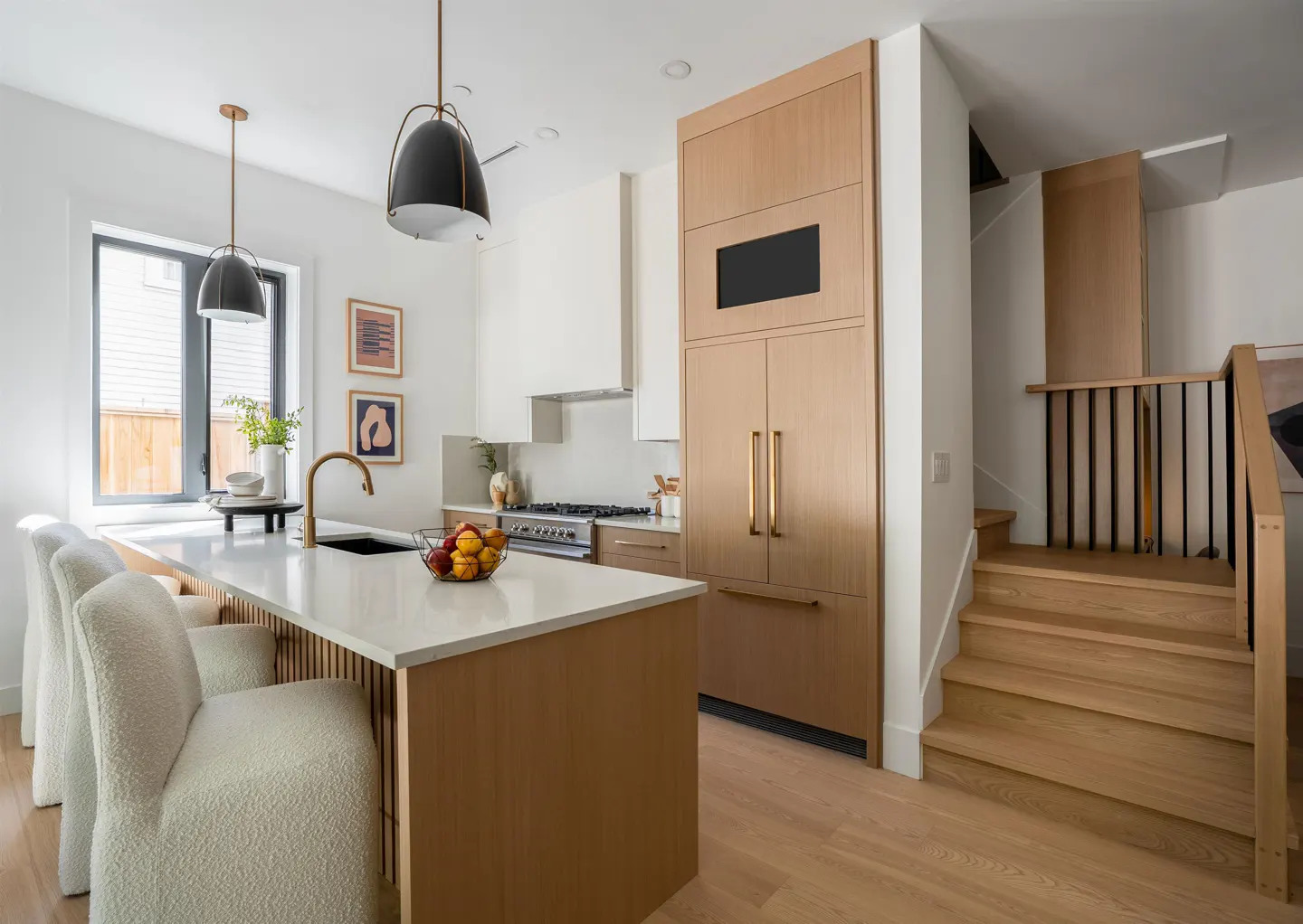 Bright kitchen with white walls, wood floors, and a large island with white countertop and seating. Stairs lead to the next floor.