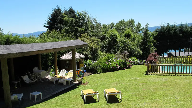 Lawn with lounge chairs, a covered patio with seating, a playhouse, and a pool surrounded by lush greenery.