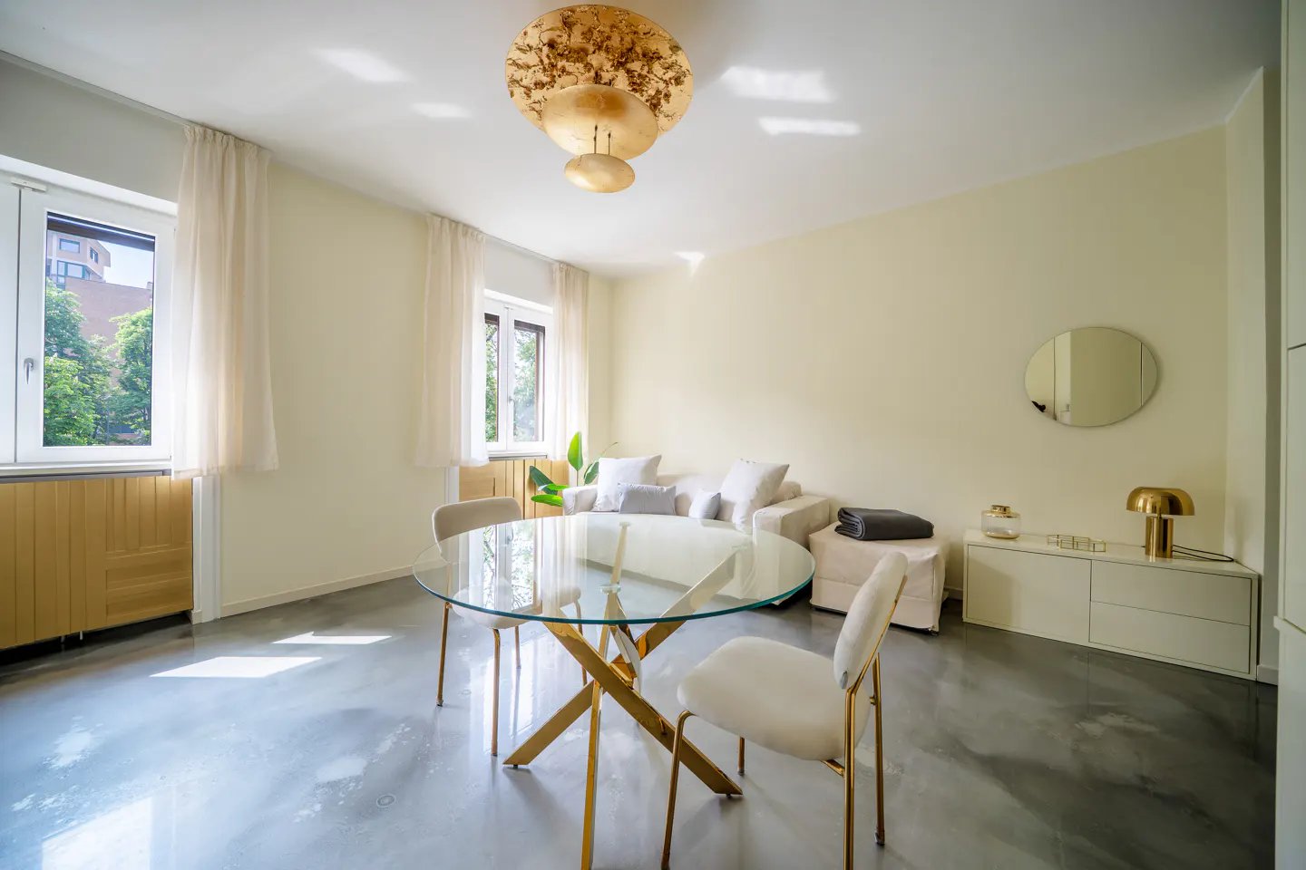 Bright living room with a glass table, gold accents, and a cream-colored sofa. Windows let in natural light.