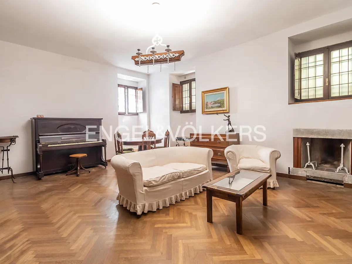 Bright living room with herringbone wood floors, white sofa and chair, piano, fireplace, and dining table.