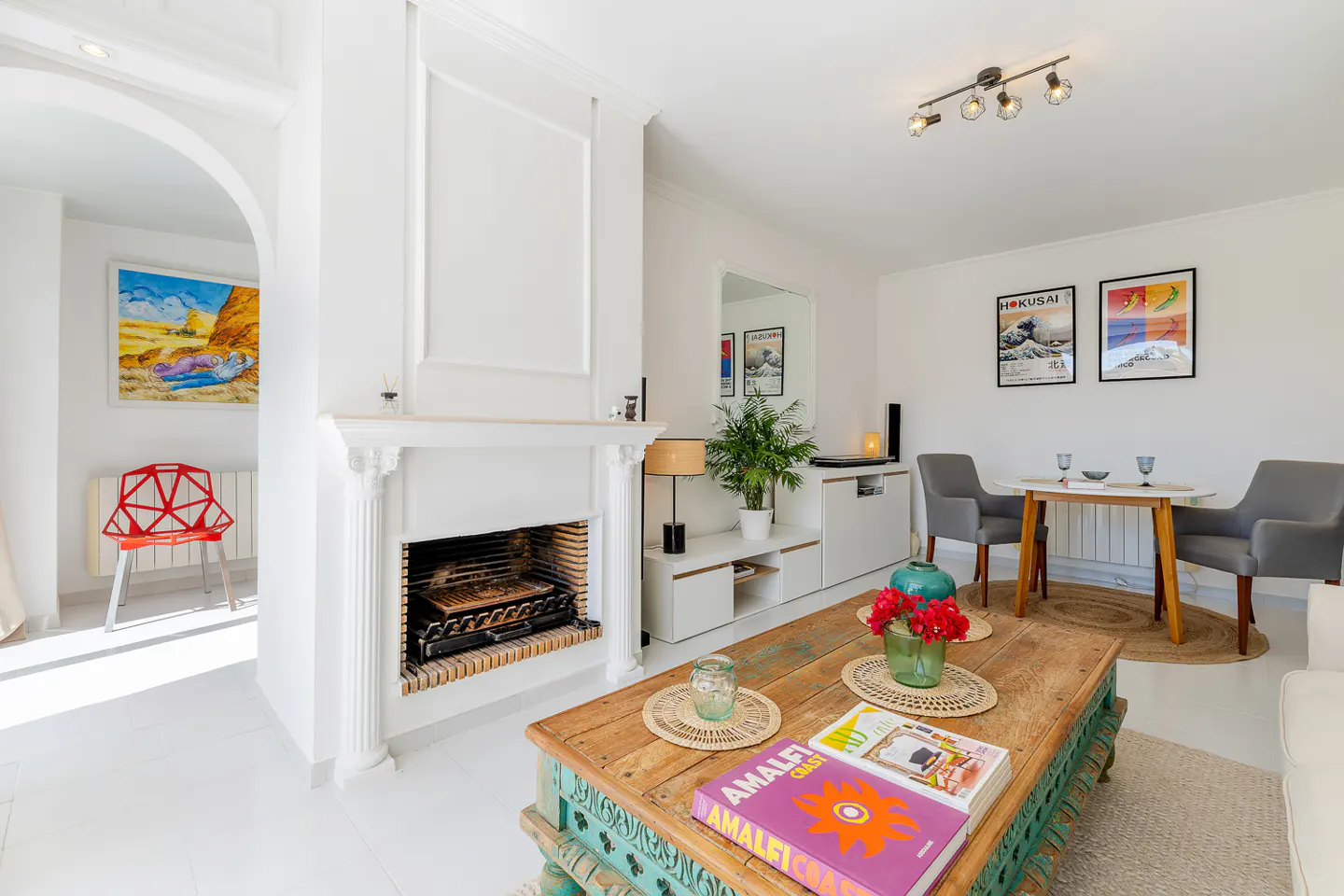 Bright living room with white walls, fireplace, and a rustic coffee table with books and flowers. Dining table with gray chairs in the background.