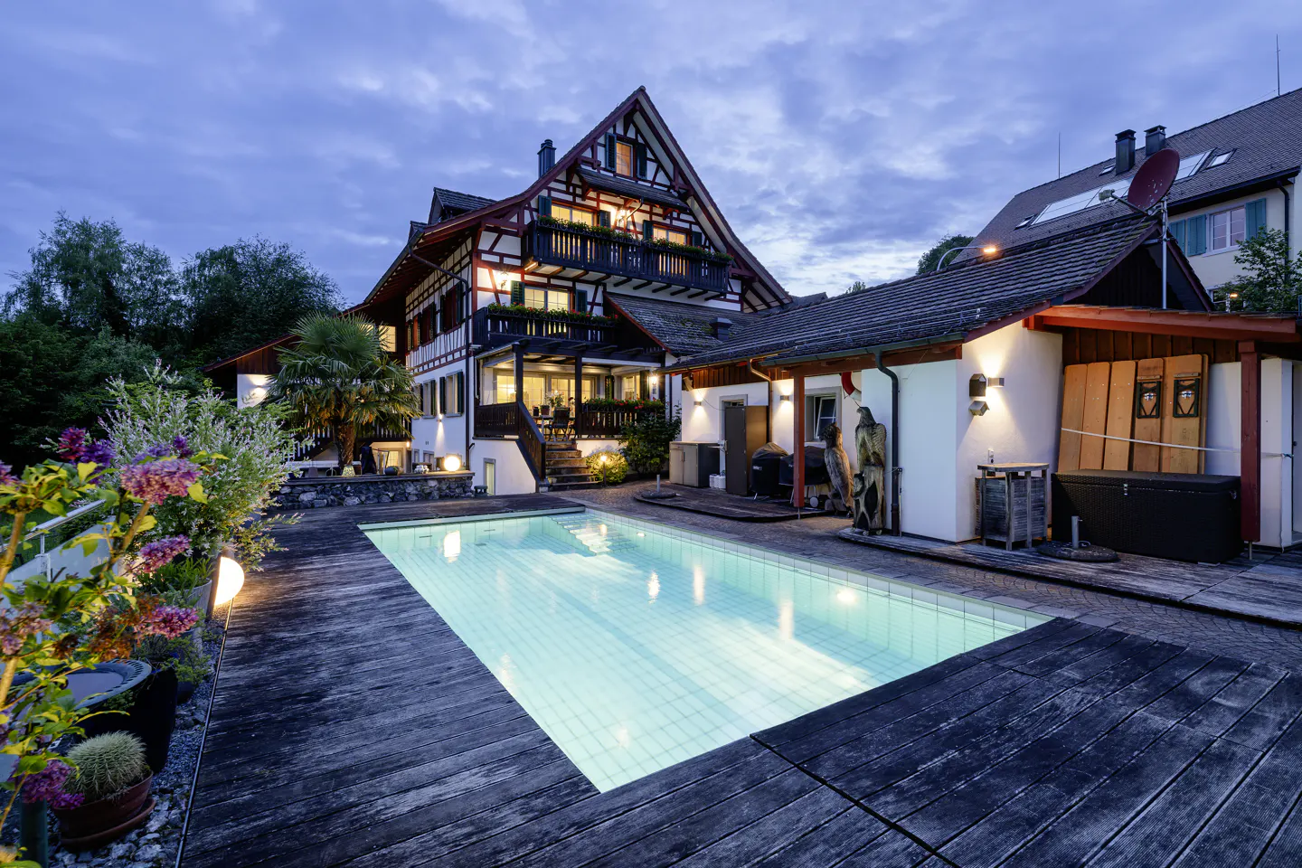 A two-story house with a pool at dusk. The house has a red and white facade with dark wood trim. The pool is surrounded by a wooden deck.