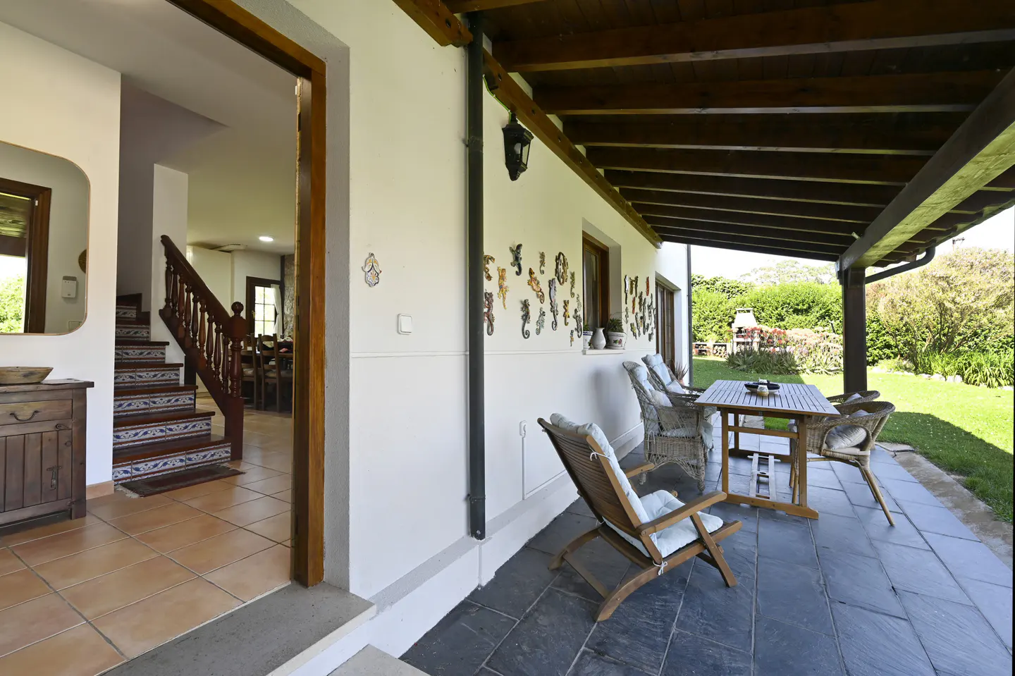 Covered patio with a table and chairs, leading to a home's interior with a staircase. The patio overlooks a green lawn.