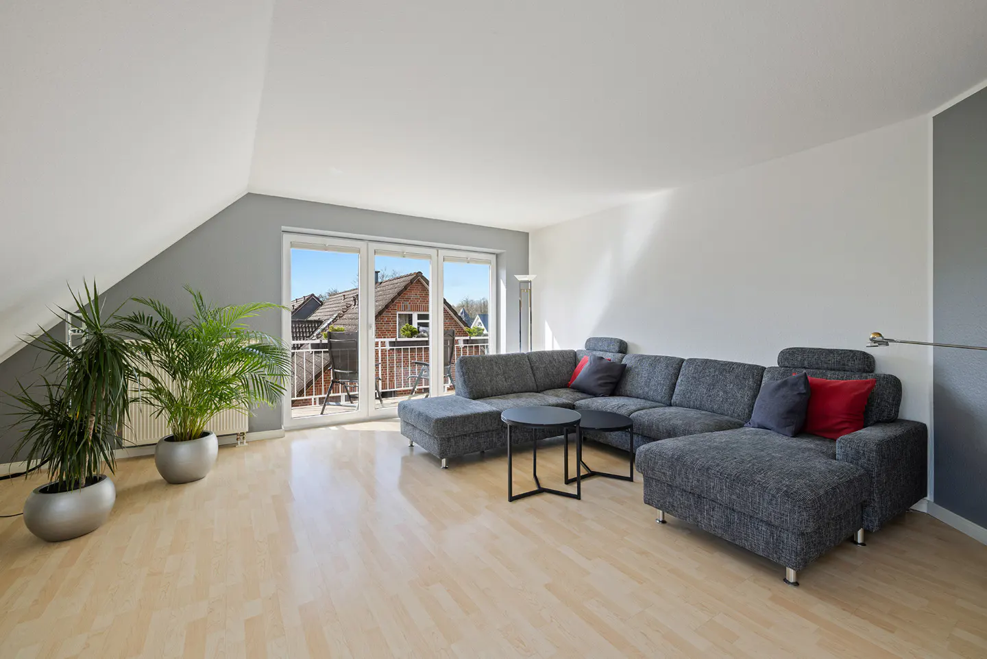 Bright living room with gray sectional sofa, black tables, and potted plants. Balcony doors open to a view of rooftops.