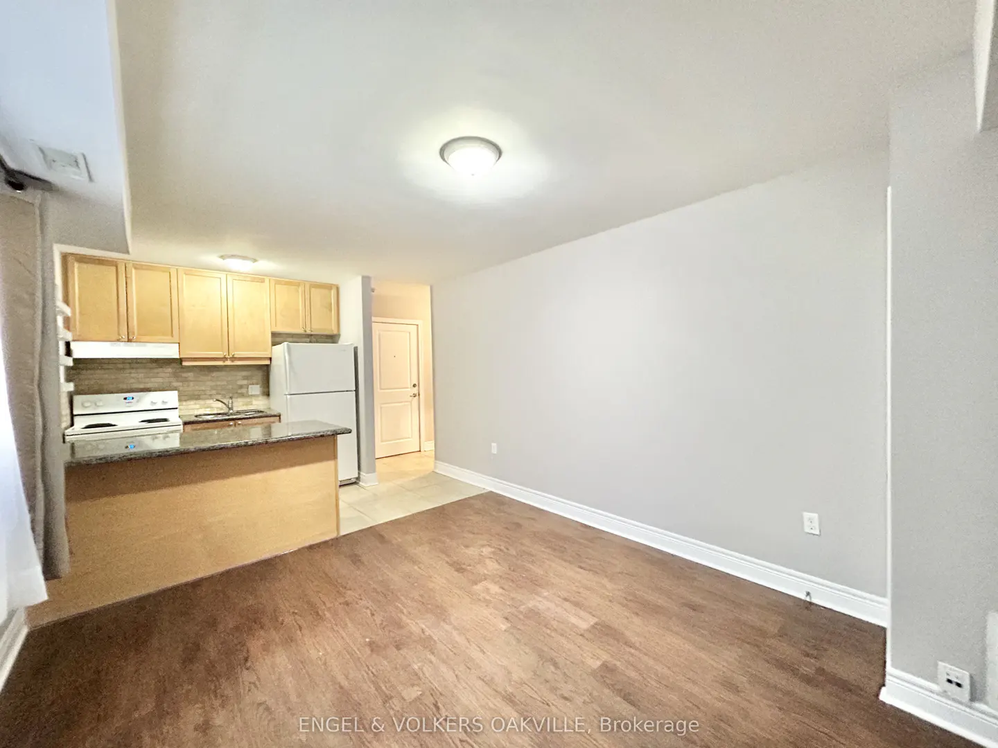A wide shot of a studio apartment with wood floors, a kitchen with light wood cabinets, and a white refrigerator.