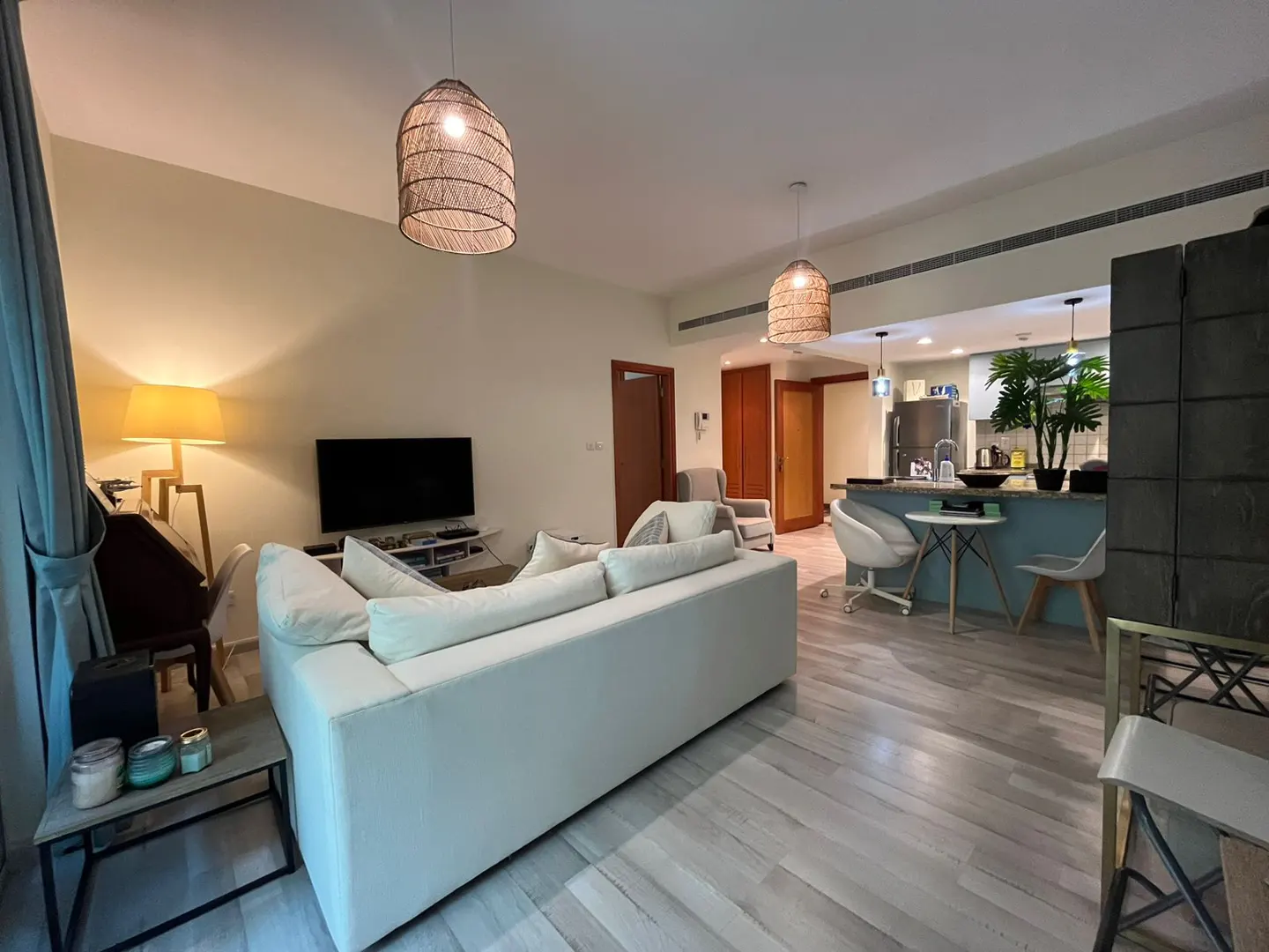 Modern open-plan living room with a white sofa, TV, and a kitchen area with a breakfast bar and woven pendant lights.