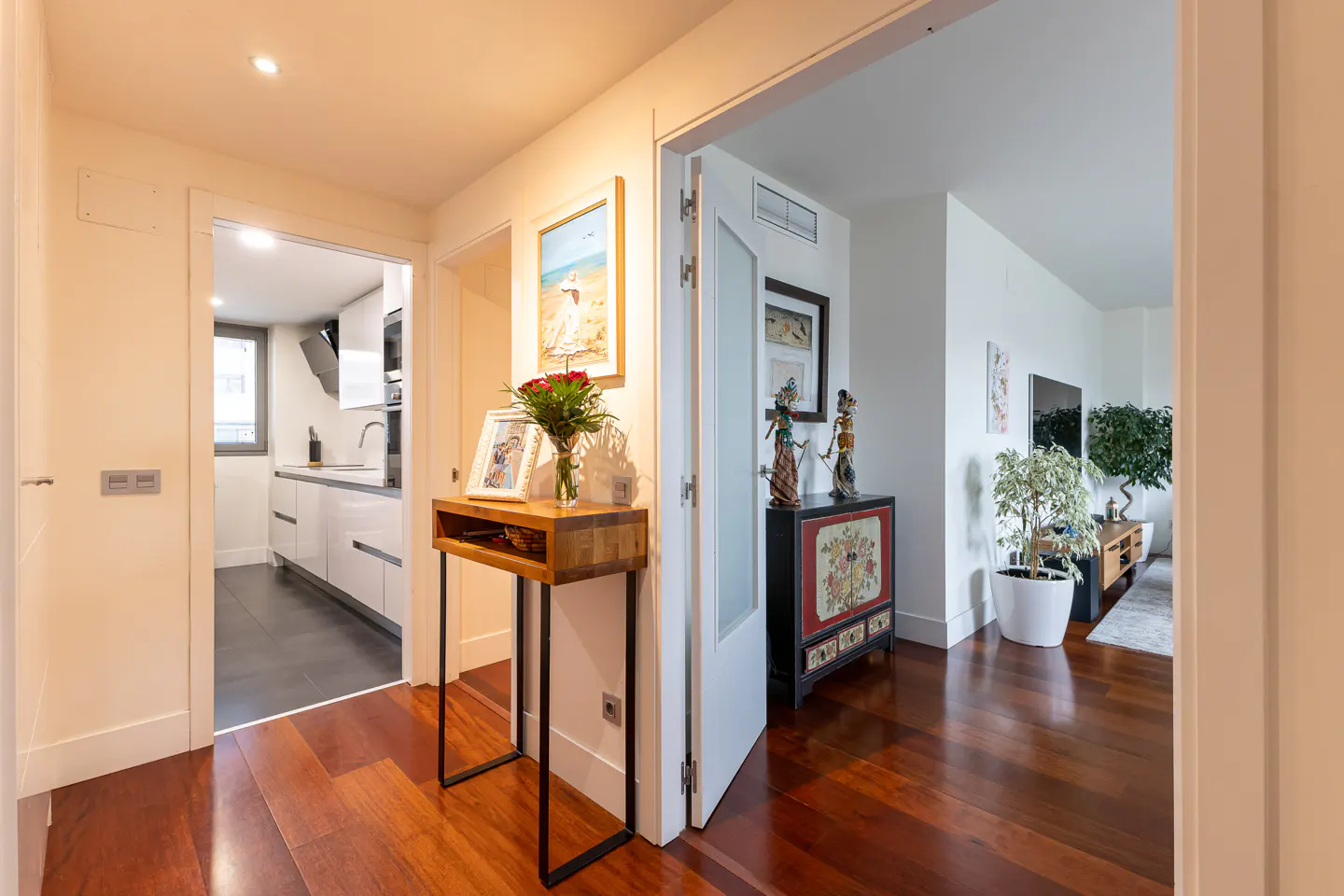 Hallway view with hardwood floors, white walls, and doorways to a kitchen and living room. A wooden table holds flowers and a framed photo.