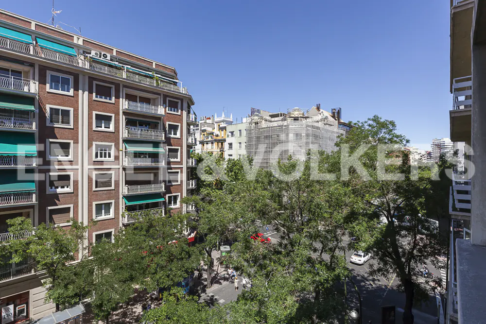 View of a city street from a balcony, featuring a brick apartment building with green awnings and lush trees below.