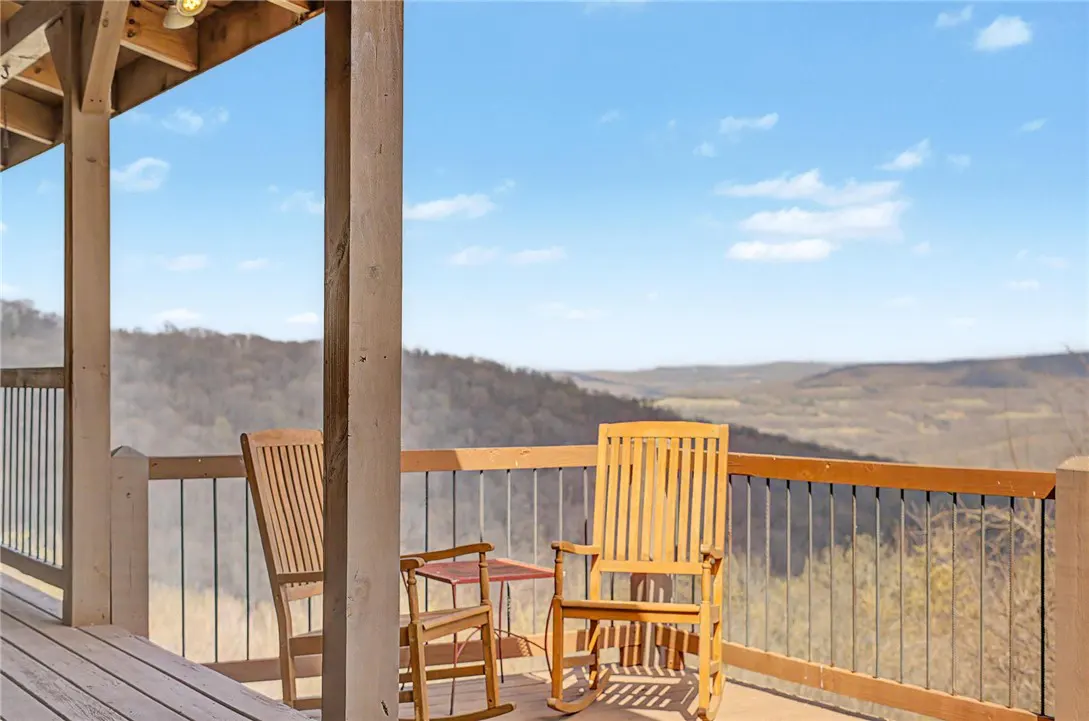 Wooden rocking chairs sit on a porch overlooking a mountain range under a blue sky with scattered clouds.