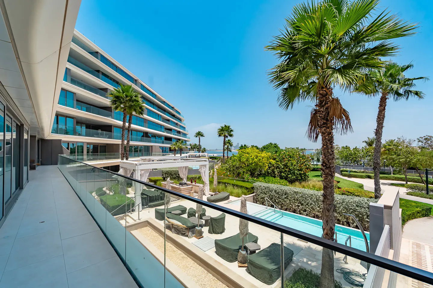 Balcony view of a modern condo building with palm trees, pool, and lounge area under a bright blue sky.