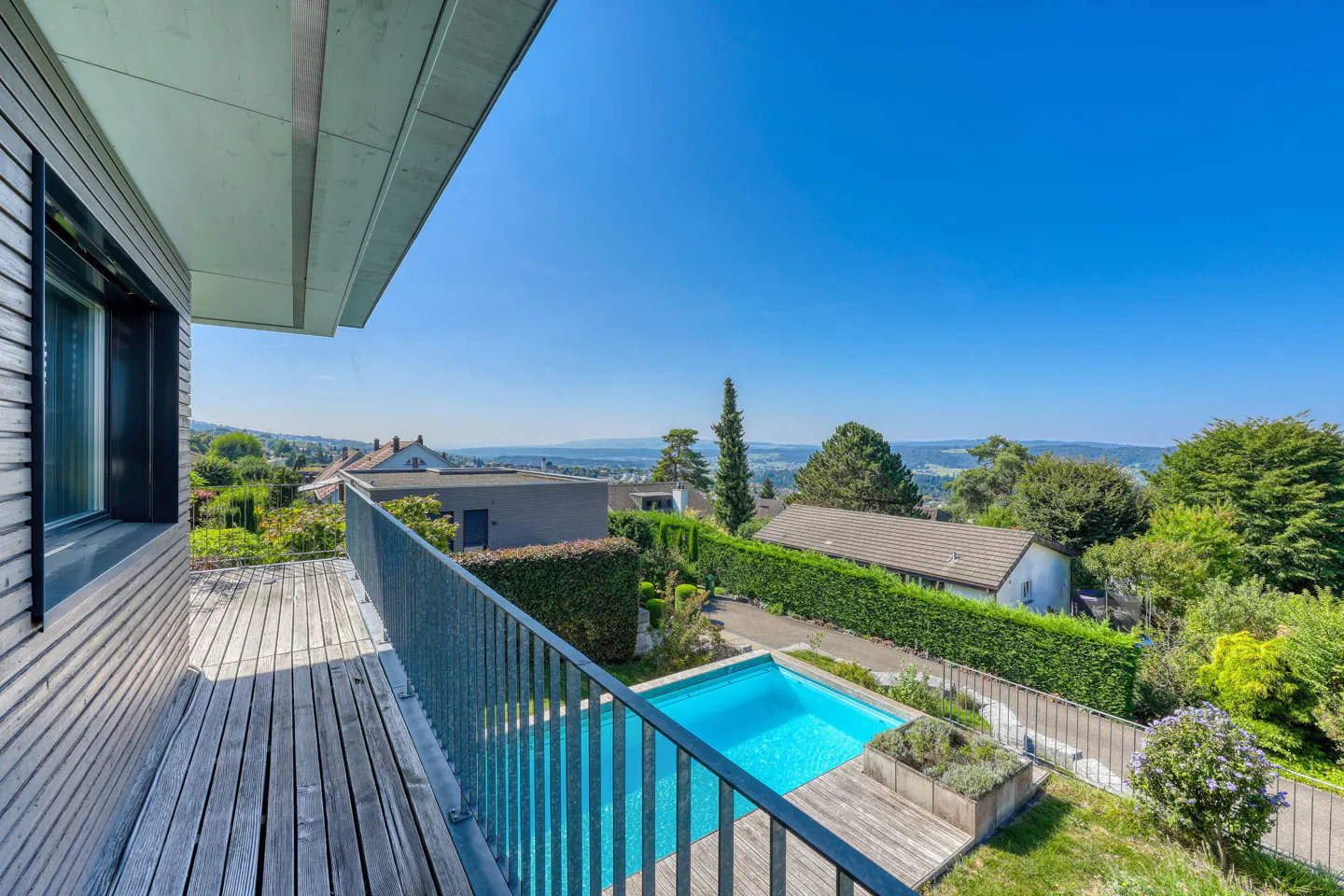 View from a wood balcony overlooking a blue pool, green hedges, and houses under a clear blue sky.