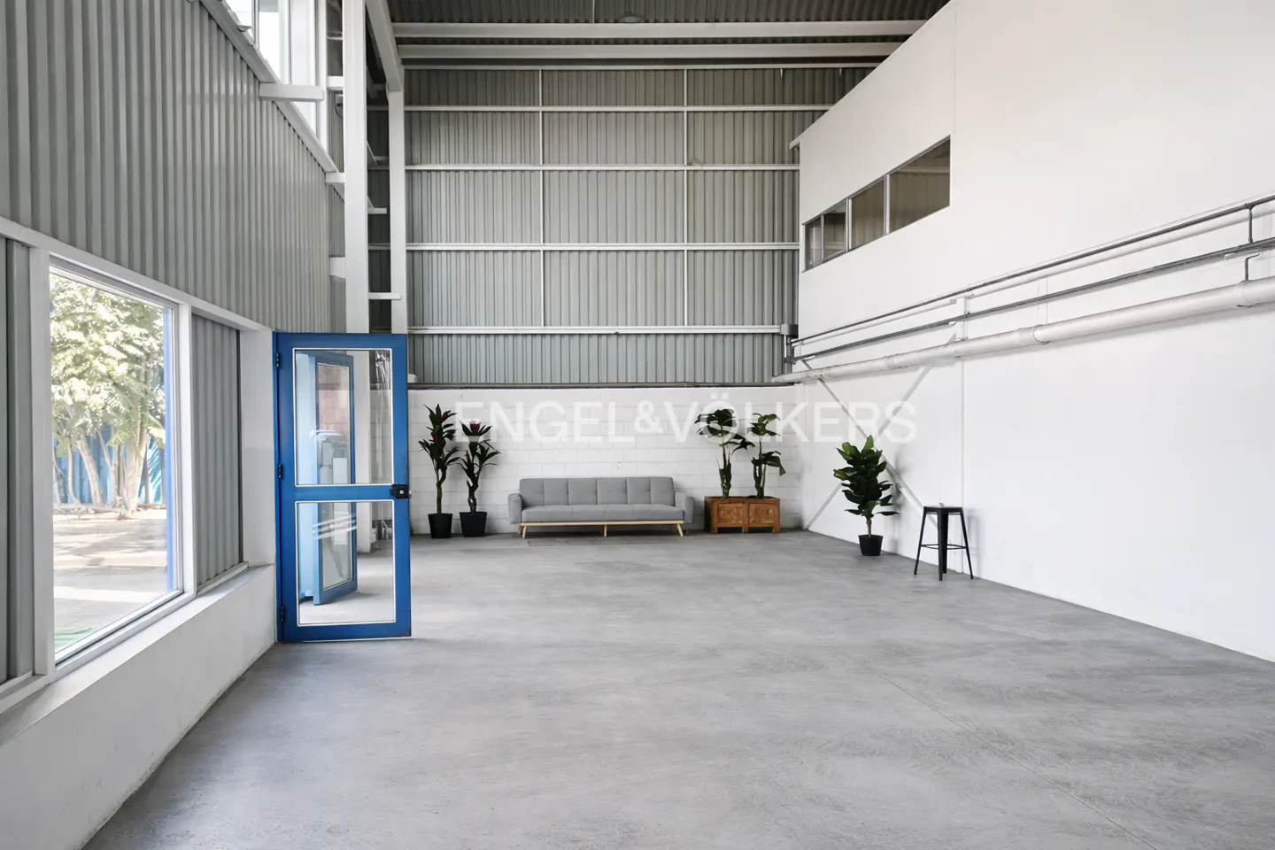 Interior of a large, empty warehouse with a gray sofa, plants, and a blue door. The walls are white and gray metal.