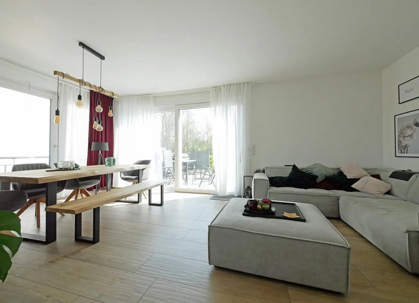 Bright living room with wood floors, a dining table with chairs and bench, and a gray sectional sofa with pillows. A sliding glass door leads to a balcony.